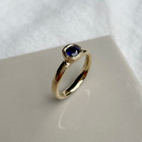 Dark Blue Sapphire cushion cut engagement ring, ethically made in 18ct yellow gold and inspired by artefacts and ancient antique jewellery | Bespoke rubover bezel engagement rings available in 9ct, 14ct, 18ct gold and platinum | Crafted from 100% recycled metals in south London with the philosophy that roughness and refinement can coexist